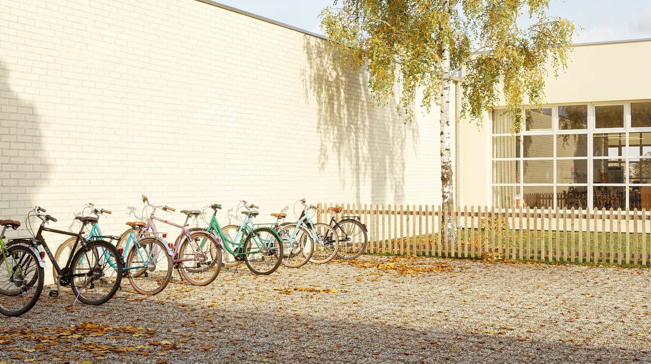 Cyklar parkerade mot en skolvägg en klar höstmorgon, med grusplan, björk och ett brett fönster